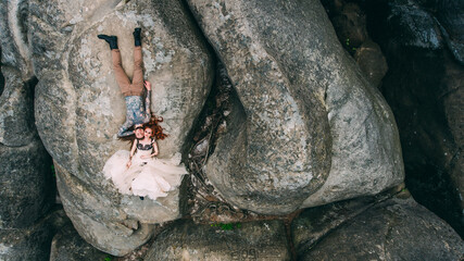 Young couple lying on the edge of a cliff around nature and circle arch. Man with long hair beard and tatto. Woman in white and black dress and long red hair. Aerial.