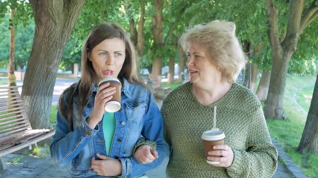 Happy elderly woman adn young woman walking park lane drinking coffee. Adult Mother and Daughter walking in the park