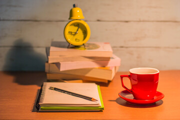Book pile for notebook, red coffee mug and yellow clock. Put on a brown desk