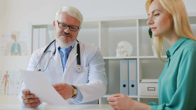 Caucasian midle aged male doctor explaining something to patient using sheet of paper sitting at desk in office.