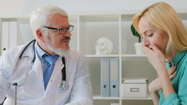 Coughing sick female patient complaining to caucasian midle aged doctor in glasses sitting in office about her throat.