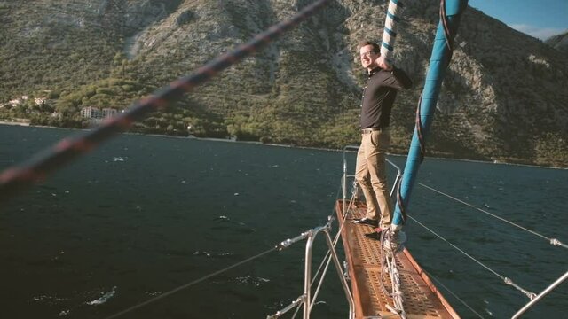 Young man stands on bowsprit of sailboat and screams outdoors. Handsome male with glasses, in black sweater and brown trousers stands on iron bow of yacht, holding rope with right hand, while left