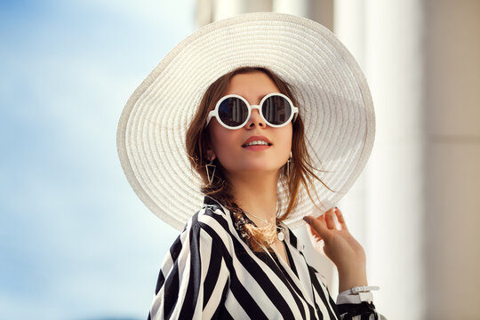 Outdoor close up portrait of young beautiful happy girl posing in street. Model wearing stylish round sunglasses, wide-brimmed hat, stripped black-white blouse. City lifestyle, female fashion concept