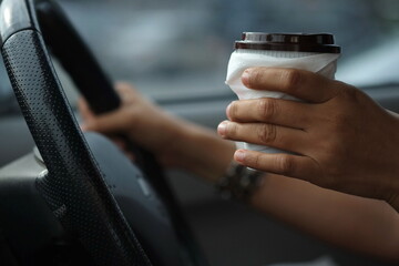 Obraz premium A woman holding a coffee mug with the other hand holding a steering wheel while driving a hurry.