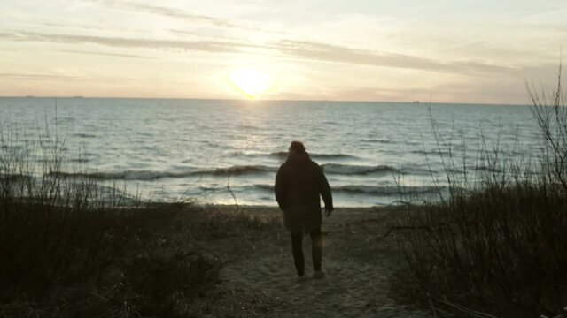 Back view of young man walking on the shore of the sea on sunset. Lonely male spending time alone in beach in cold day.