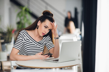 Fototapeta premium Pensive talented attractive female journalist is thinking over creative solutions for making article working in cafe, charming skilled designer noting ideas for start up in her smartphone.
