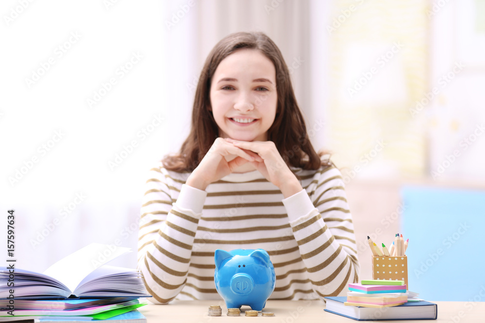 Wall mural Smiling girl sitting at table with piggy bank and stationery. Saving for education concept