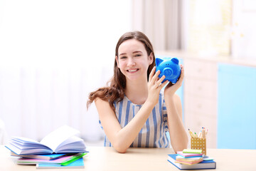 Smiling young girl sitting at table and holding piggy bank in hands. Saving for education concept