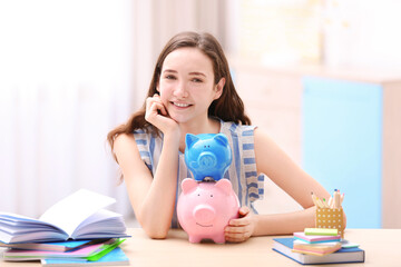 Obraz na płótnie Canvas Smiling girl sitting at table with piggy banks and stationery. Saving for education concept