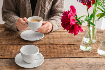 Female hands holding cup of coffee over wooden table with peonies in glass vases