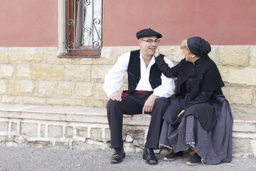 Couple dressed with one of the traditional Basque Country dresses, outdoors, seated in front of a house.