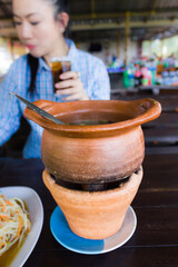 Baked clay pot of hot soup or Asian food on wooden table with background women hold glass of soda