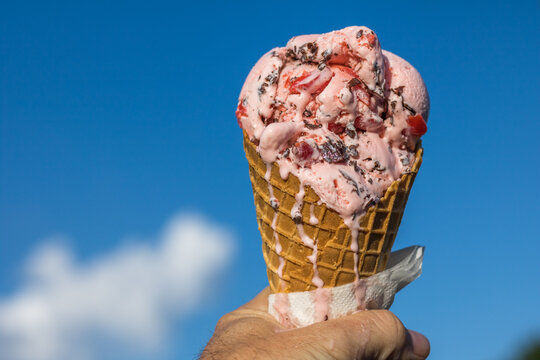 Horizontal close up of melting cherry chocolate chip ice cream in a waffle cone being held in a hand with a bright blue sky with white clouds in the background