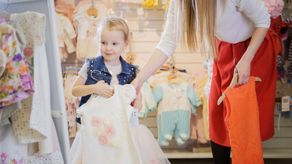 Obraz premium The girl holds in her hands hangers with dresses in front of a mirror in a children's clothing store
