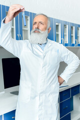 Fototapeta premium pensive senior bearded scientist in white coat looking at tube with reagent in chemical laboratory