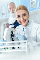 caucasian smiling scientist in white coat working with microscope and reagents while looking at camera with colleague behind in lab