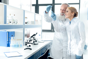 two smiling chemists in white coats looking at tube in chemical laboratory with microscopes