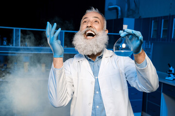 happy mature scientist in lab coat holding flask with reagent at laboratory