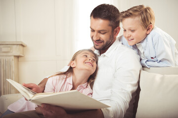 cheerful father reading book to his daughter and son while sitting on sofa