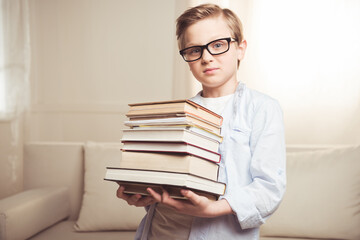confident schoolboy wearing eyeglasses and holding pile of books at home