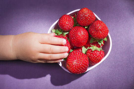 Bowl of strawberries with child hand in motion blur stealing a fruit. Top view, purple background