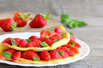 Fresh strawberry egg omelette. Omelette filled with strawberries slices and garnished with mint leaves on a plate. Fresh strawberries on a wooden table. Clean eating sweet breakfast omelette recipe