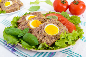 Meatloaf stuffed boiled egg with fresh tomato and lettuce leaves on a plate on a white wooden table.