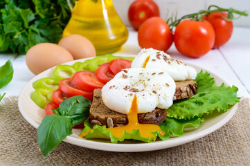 Sandwich with egg poached, lettuce, black bread with seeds, tomatoes, sweet pepper on a plate on a white wooden table.