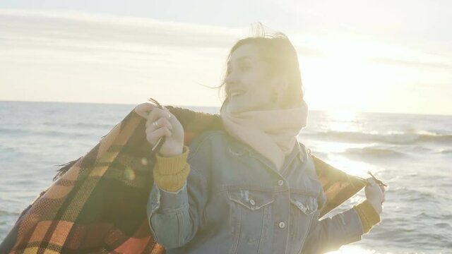 Young beautiful woman raises hands up with plaid on the shore of the sea. Happy female enjoying the sunset on the beach.