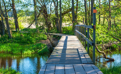 Wooden trail bridge over a gentle stream. Trail marker at the side. Some trees on the other side and after that the landscape is opening. Havang nature reserve in Sweden.
