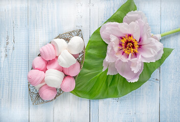 Marshmallows in a vase next to peony flower on a green leaf on a light wooden background. 