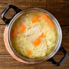 Homemade chicken soup with noodles and vegetables in metal pan on wooden table.