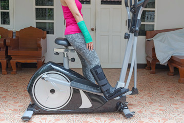 injury woman wearing sportsware with green cast on hand and arm, black splint on leg  sitting on exercise bike at home, body injury concept