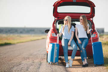 Young, slim, beautiful women, a blonde in a blue dress and a red-haired girl in a red dress,long black eyelashes, two best friends are preparing to travel on a red car with suitcases and things
