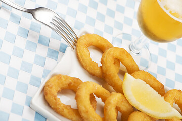 Plate of squid to the Roman next to a beer on tablecloth of white and blue squares. Food squids.