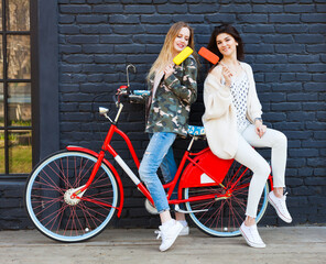 Fototapeta na wymiar Two girlfriends dressed in fashionable outfits rest, eat ice cream sitting on a trendy red vintage bicycle