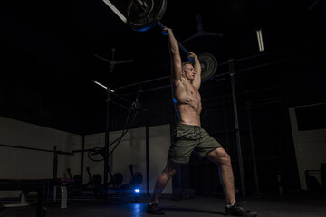 Fototapeta na wymiar Muscular White Caucasian man does a barbell overhead press in a dark grungy gym wearing shorts and showing muscular body 