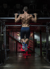 Muscular White Caucasian man performing a pull-up exercise in a dark grungy gym wearing shorts and...