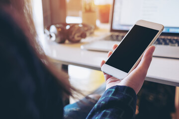 Mockup image of a woman holding white mobile phone with blank black screen while using laptop with headphone on the table in office