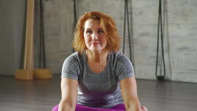 Portrait of adult woman, sitting on the rug for yoga and looking in camera. A woman of 35-40 years makes wrist-lock grip behind the back. A middle-aged woman practicing yoga, changing asanas and poses