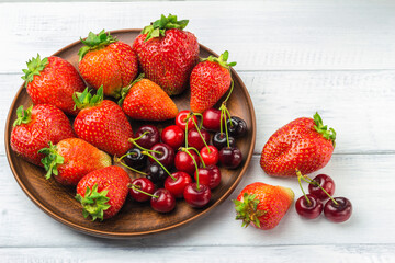 Harvest of fresh organic berry, strawberries and cherries in brown plate of clay on white wooden planks background