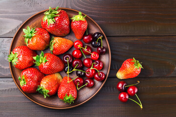 Harvest of fresh organic berry, strawberries and cherries in brown plate of clay on dark wooden planks background, top view