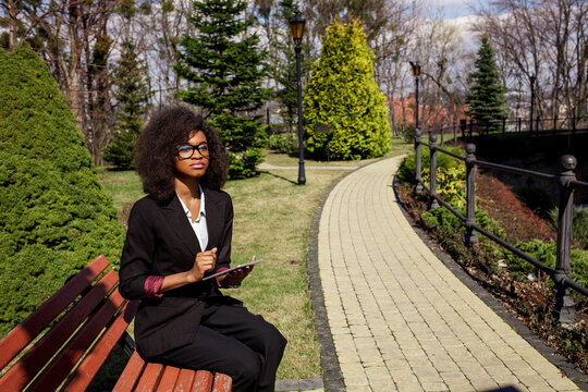 Beautiful black businesswoman sitting on the bench with lablet in her hands. Woman wearing suit and glasses.