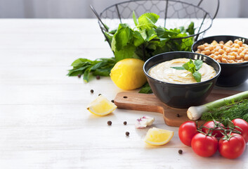 Bowl with hummus, chickpeas, lemon, cherry tomatoes and herbs on a white rustic wooden background. Middle Eastern cuisine. Background hummus with place for text.