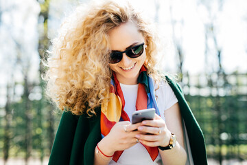 Beautiful woman with curly hair wearing sunglasses and green jacket holding smart phone in her hands browsing internet enjoying online communication while standing against green park background