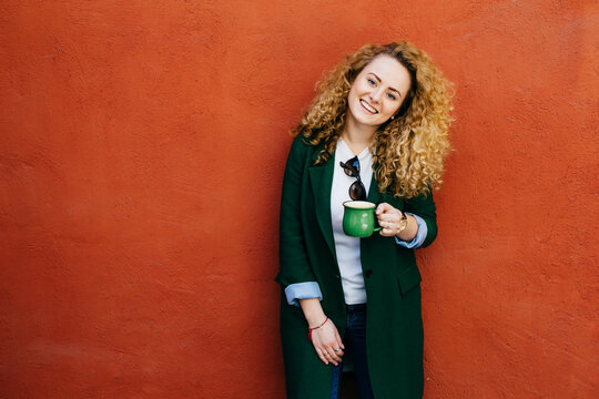 Pretty curly female wearing stylish clothes holding cup of coffee having broad smile on face looking directy into camera isolated over orange wall with copy space for your text or promotional content
