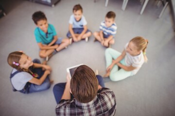 Overhead view of teacher and schoolkids sitting in library - obrazy, fototapety, plakaty