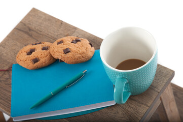 Notepad, cup, pen in turquoise color with chocolate chip cookies. wooden table and white background. Great morning and start of the day. Students accessories and student lifestyle.