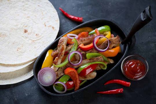 Frying pan with pork fajitas and tortillas on a dark metal background, elevated view, horizontal shot