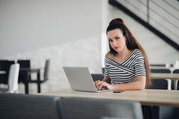 Attractive self-employed woman is working on project, using wi-fi on her laptop, sitting at wooden table with hot drink and smartphone, looking at camera with serious expression.